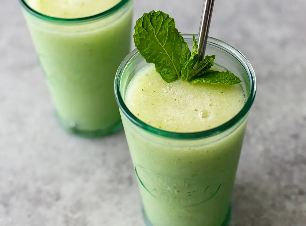 Two Cucumber Pear Mojito Slushies. Both are in glass containers, one has a metal straw, and is topped with fresh mint.