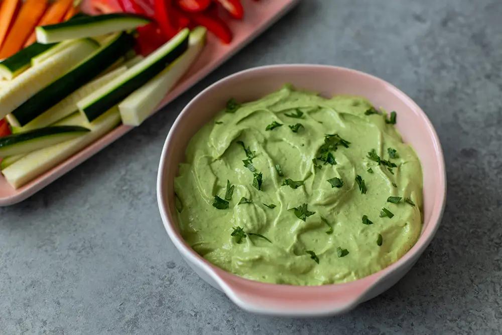 Avocado Cashew Dip next to some sliced vegetables. It is a green dip topped with fresh herbs.