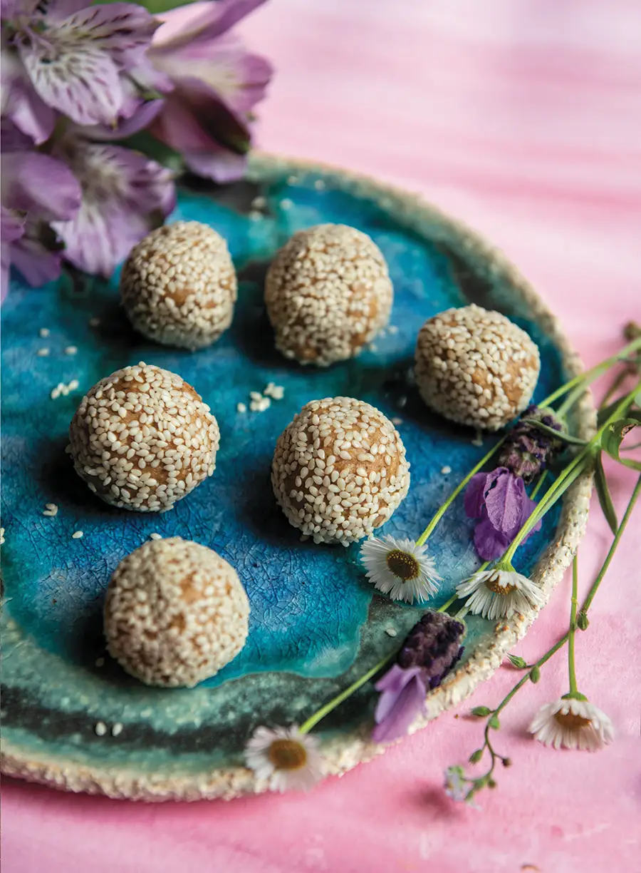 6 Tahini Snack Balls sit on a blue plate surrounded by spring flowers. Each Tahini Ball is covered in sesame seeds.